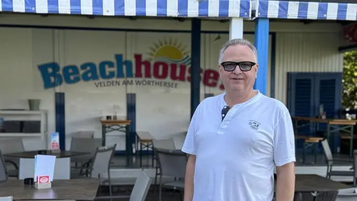 Ein Mann mit Brille steht in einem Gastgarten, im Hintergrund ist das Logo „Beach House“  zu sehen