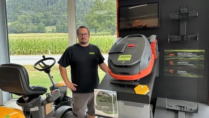 Ein Mann steht neben einem Rasenmäherroboter. Neben ihm befindet sich ein Mähtraktor.