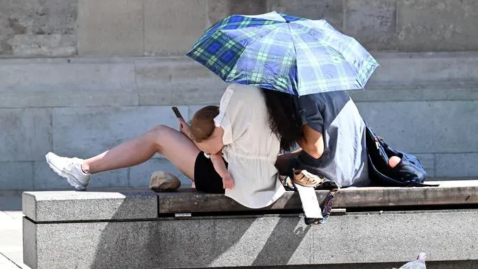 Das Bild zeigt zwei Menschen, die unter einem Regenschirm zusammengekauert auf einer Bank in der Sonne sitzen