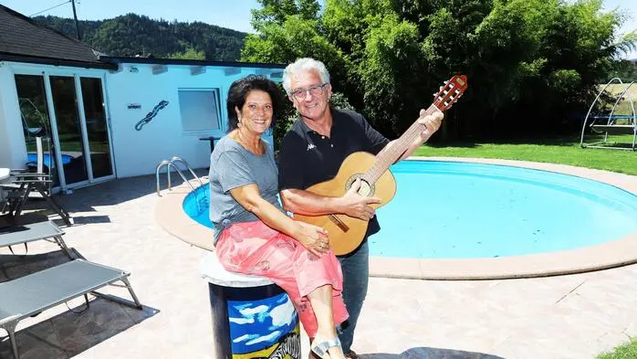Ein Mann und eine Frau bei Sonnenschein, sitzend vor einem Pool. Der Mann hält eine Gitarre in den Händen.