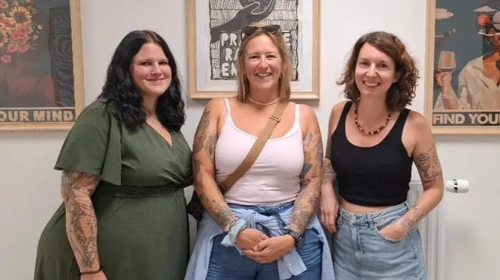 Drei Frauen stehen vor einer Wand mit bunten Bildern und lächeln in die Kamera