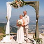 Ein Brautpaar steht unter einem Hochzeitsbogen, im Hintergrund sind Berge zu sehen