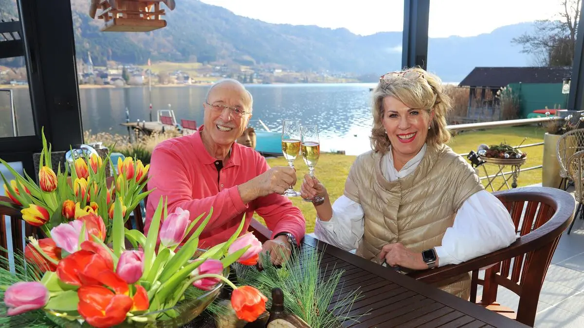 Ein älterer Herr und eine Dame sitzen an einem Tisch, im Hintergrund ist ein See, der Tag ist sonnig