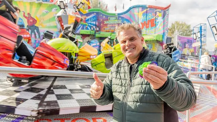 Ein Anfang 50-jähriger Mann in einer grünen Jacke hält Jetons in der Hand, macht einen Daumen hoch und lächelt. Im Hintergrund befindet sich ein Fahrgeschäft in bunten Farben.