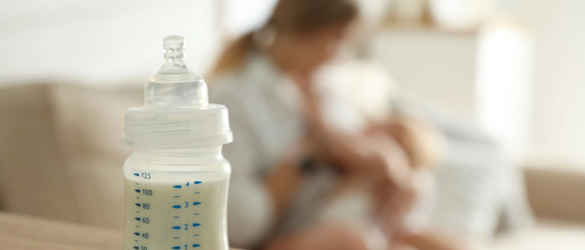 Babyfläschchen im Vordergrund, eine Frau mit Baby im Arm im Hintergrund