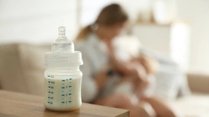 Babyfläschchen im Vordergrund, eine Frau mit Baby im Arm im Hintergrund