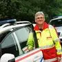 Ein Mann mit Brille und grauen Haaren lacht in Einsatzkleidung vor einem Auto in die Kamera