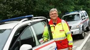 Ein Mann mit Brille und grauen Haaren lacht in Einsatzkleidung vor einem Auto in die Kamera
