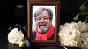 Ein Bild von einem Mann mit Brille und Bart in einem braunen Rahmen; daneben liegen weiße Blumen
