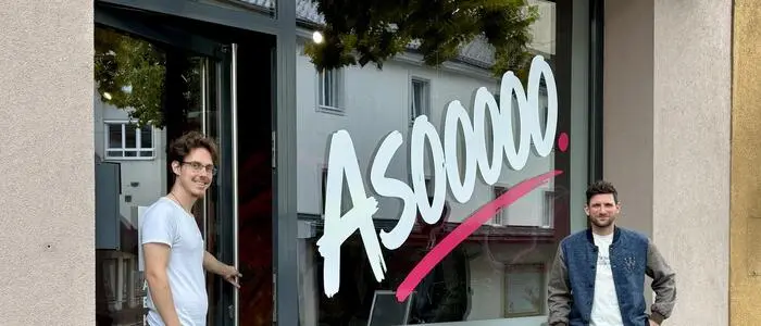 Zwei Männer stehen vor einem Geschäft auf dem „Asoooo“ steht