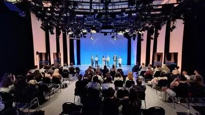 Ein Studioraum mit Lichtern an der Decke, im Vordergrund sitzen Jugendliche, im Hintergrund stehen sechs Erwachsene vor einem Blauen Hintergrund