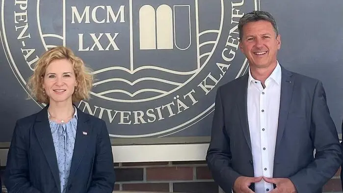 Man sieht trotz Sparkurses Eva-Maria Holzleitner und Daniel Fellner lachend vor dem Wappen der Uni Klagenfurt