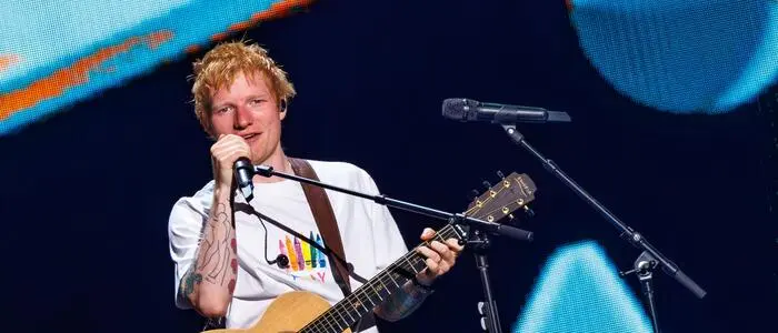 Ed Sheeran hält ein Mikro in der rechten Hand und hat eine Gitarre umgehängt.