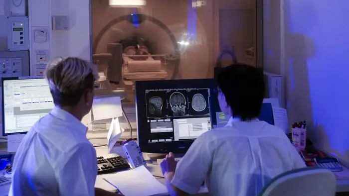 Zwei Ärztinnen sitzen vor einem Bildschirm, im Hintergrund wird eine Computertomographie einer Person gemacht.