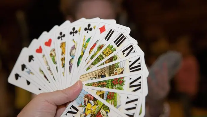 Eine Hand hält 16 Karten, dahinter sieht man unscharf eine Mitspielerin, ebenfalls mit Karten in der Hand