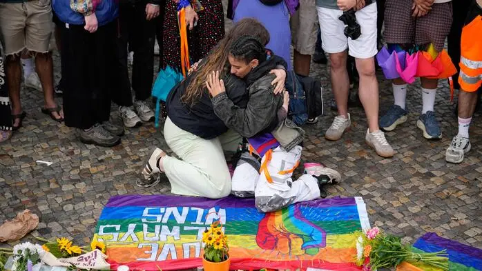 Zwei Menschen knien vor abgelegten Blumen, Kerzen und Regenbogenfarben und umarmen sich