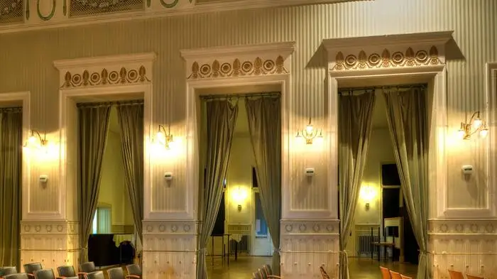 Ein Kleinod von einem Theater mit seiner Guckkastenbühne, den historischen Lustern, Bodenfliesen und Garderoben: Otto Wagner entwarf den Lageplan der Gesamtanlage auf der Baumgartner Höhe in Wien-Penzing, errichtet wurde es 1904–1907 von Franz Berger. Das einstige „Gesellschaftshaus“ steht unter Denkmalschutz, die Festwochen bespielen das Jugendstiltheater erneut mit fünf Produktionen; viele mit historischem Bezug. Denn auf der Wiese davor befindet sich das Mahnmal aus 772 Leuchtstelen, die an die Opfer erinnern sollen, die zwischen 1940 und 1945 in der NS-Euthanasieanstalt „Am Spiegelgrund“ ermordet wurden.