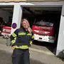 Eine blonde Feuerwehrfrau in Uniform steht vor einem Rüsthaus. Im Hintergrund sind zwei Feuerwehrfahrzeuge