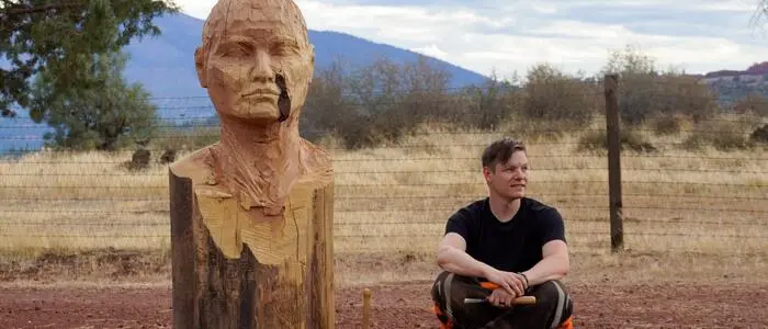 Ein Anfang 30-Jähriger sitzt am Boden. Der Untergrund besteht aus rotem Sand. Neben ihm ist eine menschengroße Skulptur aus Holz.