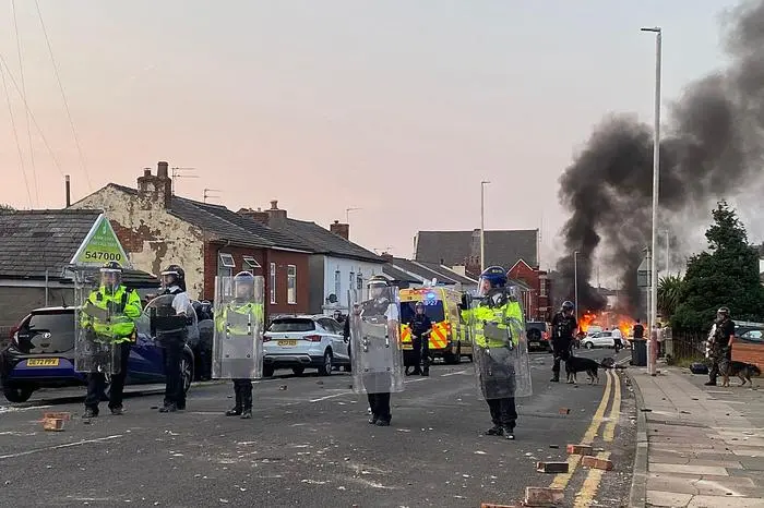 Smoke billows from a fire started by protesters as riot police stand guard after disturbances near the Southport Islamic Society Mosque in Southport, northwest England, on July 30, 2024, a day after a deadly child knife attack. Violent clashes broke out in the northern England town where a knife attack claimed the lives of three children, with around 100 protesters lighting fires and battling police. A 17-year-old male suspect from a nearby village arrested shortly after the incident remained in custody, police added, as they warned against speculating about his identity or details of the investigation. (Photo by Roland LLOYD PARRY / AFP)