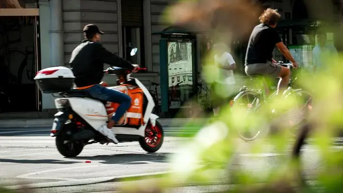 Ein Essenszusteller fährt auf seinem E-Moped auf einem Radweg, vor ihm ist ein Radfahrer unterwegs