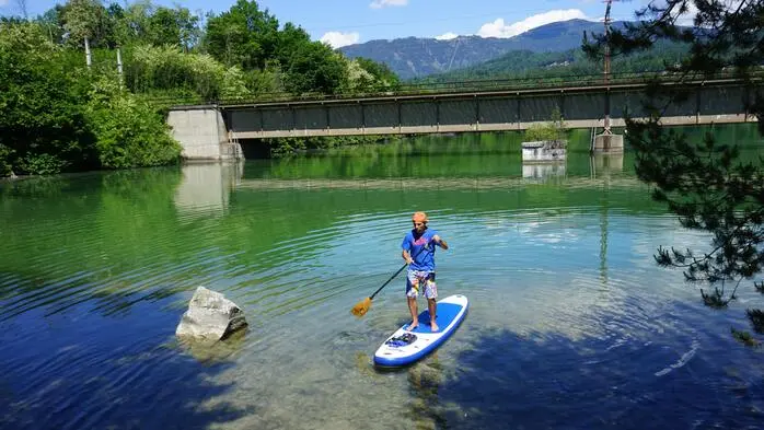 Ein Mann steht in einem Fluss auf einem SUP, im Hintergrund sieht man eine Brücke und viele Bäume