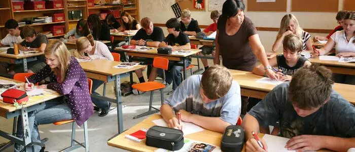 Blick in ein Klassenzimmer, Schüler sitzen schreibend an ihren Plätzen, eine Lehrerin gibt Unterstützung