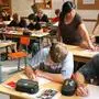 Blick in ein Klassenzimmer, Schüler sitzen schreibend an ihren Plätzen, eine Lehrerin gibt Unterstützung