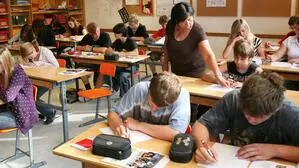 Blick in ein Klassenzimmer, Schüler sitzen schreibend an ihren Plätzen, eine Lehrerin gibt Unterstützung