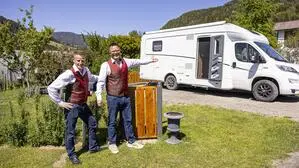 Zwei Männer mittleren Alters stehen in traditionellem Gewand vor einem Campingwagen