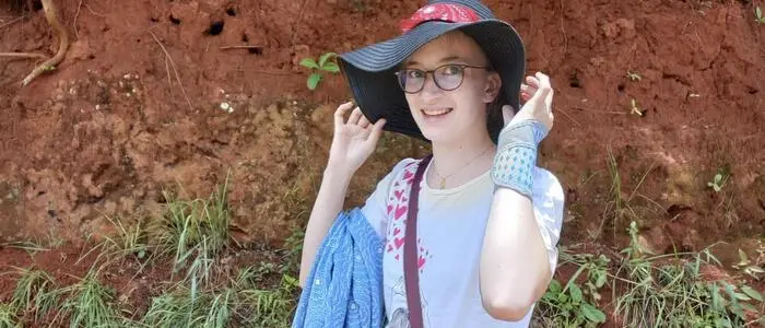 Eine junge Frau mit Brille blickt in die Kamera und hält ihren Hut mit den Händen fest