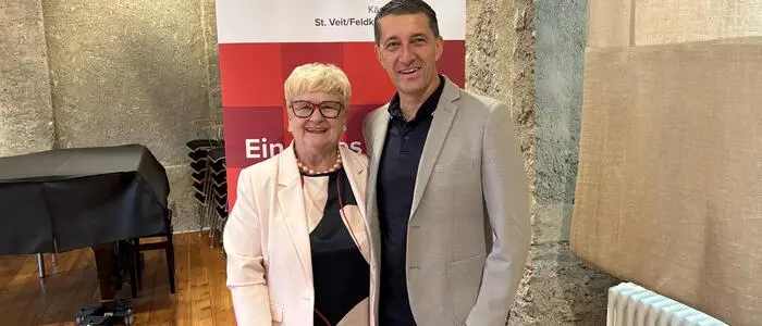 Zwei ältere Personen stehen vor einem Aufsteller und lachen in die Kamera