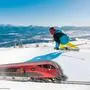 Eine Fotomontage: Ein Skifahrer bei strahlend blauem Himmel auf der Piste, darunter ein roter Zug auf Schienen | Eine Stunde und 41 Minuten dauert die Anreise vom Grazer Hauptbahnhof zum Bahnhof in Annenheim, dem Startpunkt der Gondel auf die Gerlitzen
