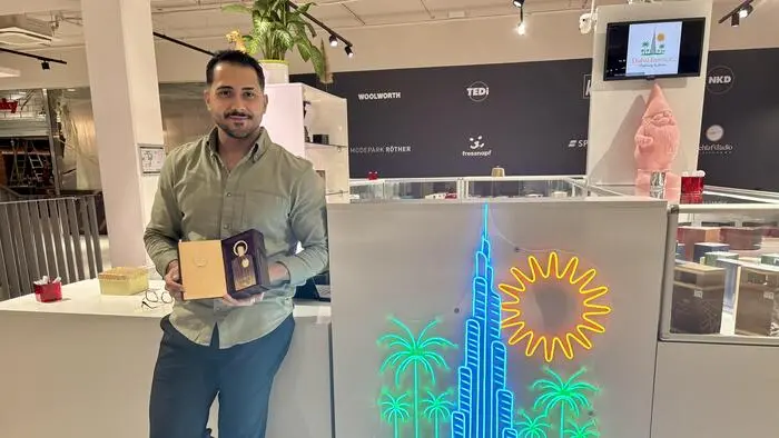 Ein Mann lehnt an einer Verkaufstheke und hält eine Parfumverpackung in den Händen. Neben ihm sieht man das Logo seiner Firma | Jiwan Mohamed lebt in Villach und möchte die weltweit bekannten Düfte aus Dubai den österreichischen Kunden näherbringen