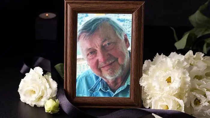 Ein Foto von einem Mann in einem braunen Bilderrahmen, daneben liegen weiße Blumen und eine schwarze Schleife