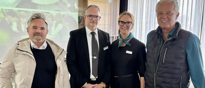 Drei Männer und eine Frau stehen vor einem Rollboard und lächeln in die Kamera