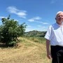 Ein weißhaariger Mann in einem weißen Hemd steht auf einer Wiese. Im Hintergrund sieht man einen Strauch.