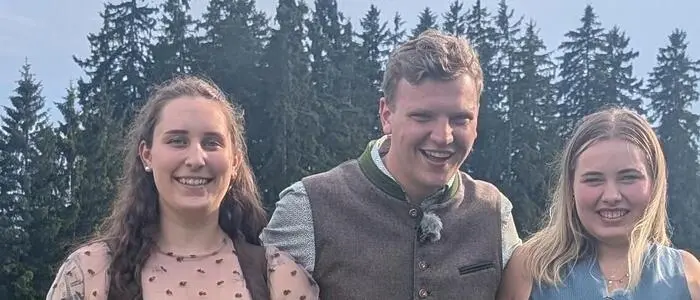 Ein Mann und zwei Frauen in Tracht stehen dicht beieinander vor einem Wald