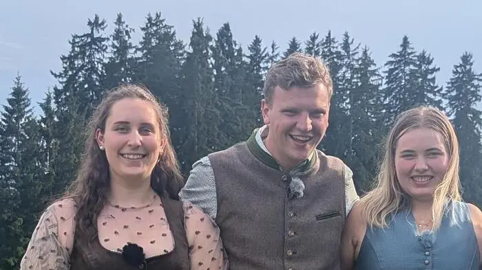 Ein Mann und zwei Frauen in Tracht stehen dicht beieinander vor einem Wald