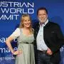 Arnold Schwarzenegger und Maria Hauser stehen lächelnd vor einer blauen Eventwand mit der Aufschrift „Austrian World Summit“. Beide posieren eng nebeneinander für ein Foto. | Arnold Schwarzenegger läutete am Donnerstag im „Stanglwirt“ den Kitz-Society-Trubel ein. Hausherrin Maria Hauser strahlte mit Babybauch