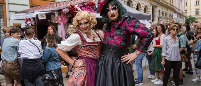 Die beiden Dragqueens stehen stark geschminkt und in rosaroten und pinken Dirndln vor der rosarot geschmückten Hütte