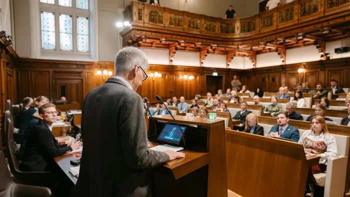 Mann mit weißen Haaren und Brille steht am Rednerpult, vor ihm sitzen die Gemeinderäte auf ihren Plätzen im Sitzungssaal