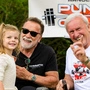 Arnold Schwarzenegger trägt eine schwarze Sonnenbrille, gemeinsam mit einem anderen Mann und einem kleinen Mädchen lacht er in die Kamera.