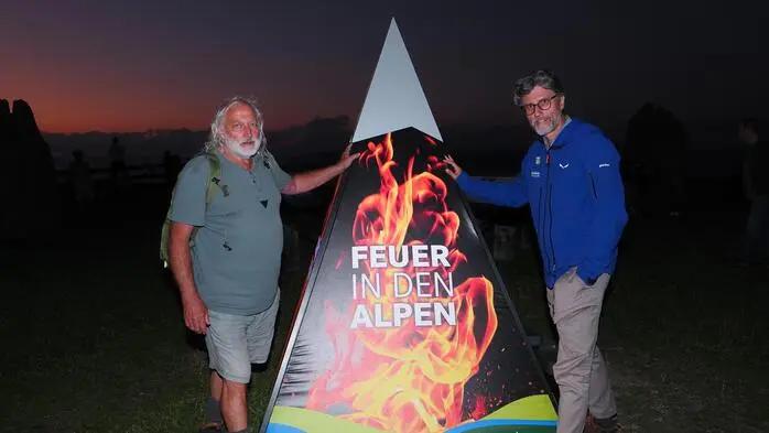 Zwei Männer stehen vor einer Kartonpyramide auf der ein loderndes Feuer abgebildet ist