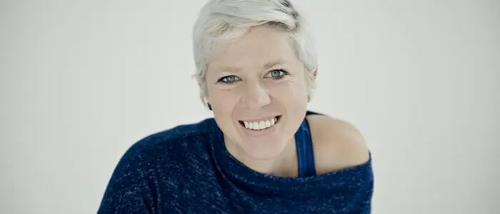 Porträt einer Frau mit kurzen hellblonden Haaren und einem breiten Lächeln. Sie trägt einen dunkelblauen Pullover.