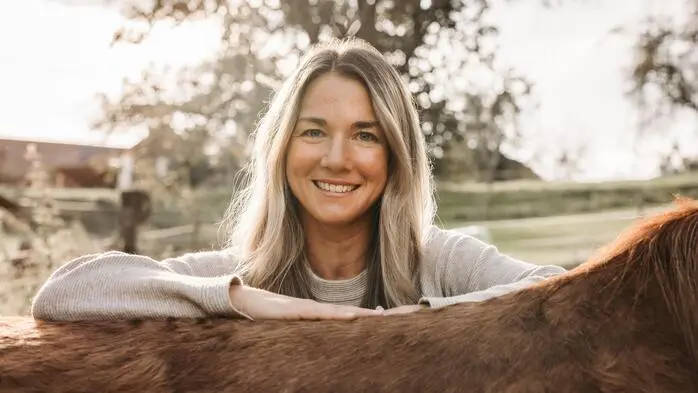 Eine blonde Frau lehnt sich an einem Pferd an und lacht in die Kamera. Der Hintergrund ist verschwommen und das Licht ist weich.