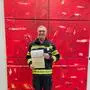 Ein Mann in Feuerwehreinsatzkleidung hält eine Urkunde in den Händen und lächelt in die Kamera.