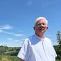 Ein weißhaariger Mann in einem weißen Hemd steht auf einer Wiese. Im Hintergrund sieht man einen Strauch.