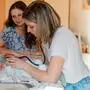 Ein Baby liegt auf einem Bett. Neben ihr steht eine blonde Frau mit einem Stethoskop. Im Hintergrund sieht man eine weitere Frau, die das Baby verliebt anschaut.
