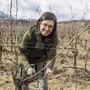Eine Frau schneidet sorgfältig die Weinreben und lächelt dabei in die Kamera.